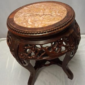 May include: An antique dark brown wooden plant stand with a round marble top. The stand has carved floral and bird designs on the sides and legs. The marble top has a mottled beige and brown pattern. The stand is approximately 40 cm in diameter.