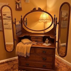May include: A wooden vanity with a large oval central mirror and two adjustable side mirrors. The piece features three main drawers with metal pulls, two smaller upper drawers, and a spindle gallery. A tan blanket and a small figurine sit on top.
