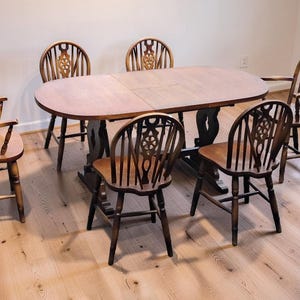 May include: A wooden dining set comprising an oval table and six chairs. The table has a light brown surface and a dark brown base. The chairs feature spindle backs with decorative carvings. The set is displayed on a light wood floor.