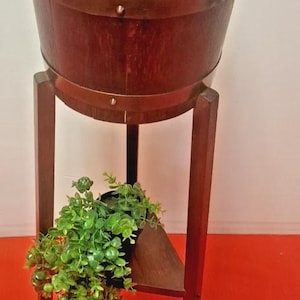 May include: A dark brown wooden planter on a matching stand. The planter has metal bands and holds a small green plant. The stand has a triangular shelf and four legs. The background is a white wall and a red surface.