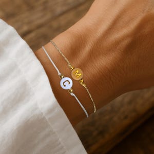 Peut inclure: Deux bracelets délicats avec des breloques personnalisées avec des lettres. L'un des bracelets est composé d'un cordon blanc avec un charm rond affichant la lettre "C". L'autre bracelet a une chaîne dorée avec un charm rond affichant la lettre "M".