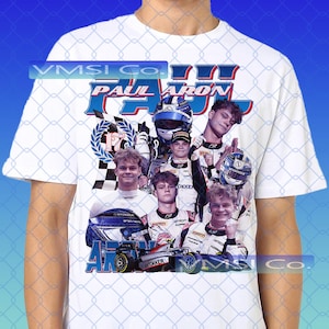 May include: White t-shirt featuring a graphic of a race car driver with the text "PAUL ARON" in blue and red. The design includes images of the driver, a race car, and a checkered flag.
