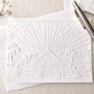 May include: White embossed Christmas cards featuring a nativity scene with a starburst design. The cards depict a stable, figures, and palm trees. Includes envelopes and a pen.