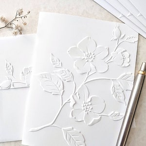 May include: White card with an embossed floral design. The design features a branch with leaves and flowers. The flowers are white and have a raised center.