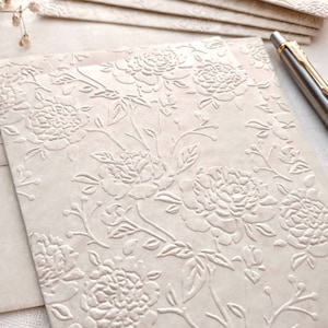 May include: A stack of off-white envelopes with a raised floral pattern. The pattern features white flowers and leaves on a cream background.