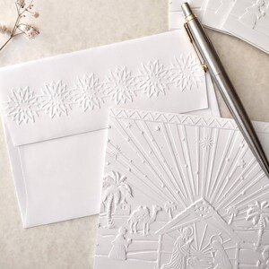 May include: White embossed Christmas cards and envelopes. The cards feature a nativity scene with palm trees, stars, and radiating light. The envelopes have a snowflake border. A silver pen with a gold clip rests on the cards.