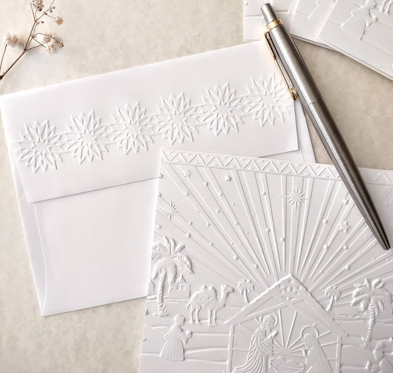 May include: White embossed Christmas cards and envelopes. The cards feature a nativity scene with palm trees, stars, and radiating light. The envelopes have a snowflake border. A silver pen with a gold clip rests on the cards.