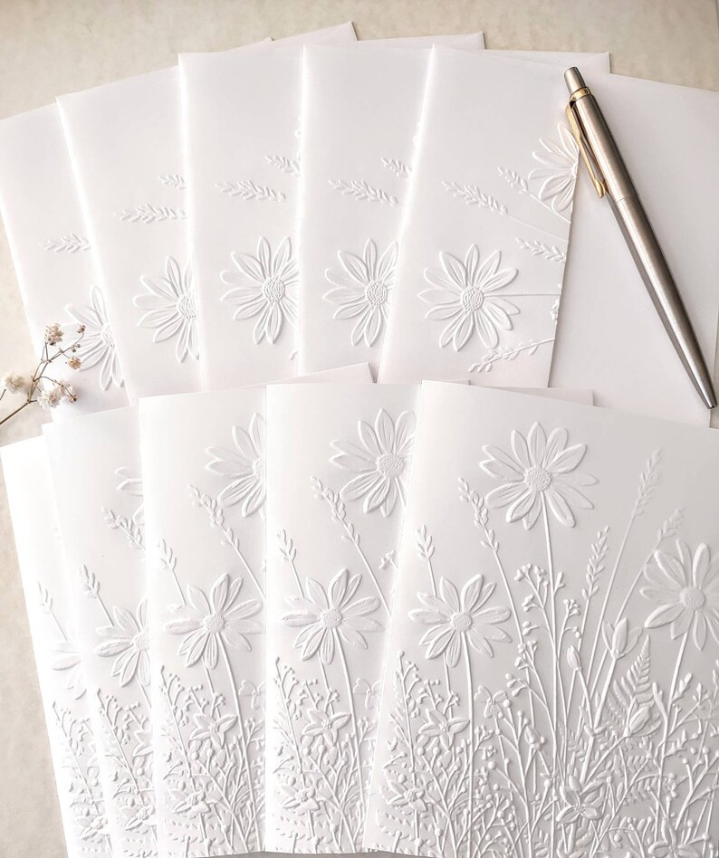 May include: A stack of white blank greeting cards with an embossed floral design. The cards feature a raised pattern of daisies and foliage.