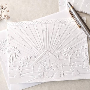 May include: White embossed Christmas card featuring a nativity scene with a starburst design. The card depicts figures, palm trees, camels, and a stable. A silver pen rests on the card, alongside a white envelope and a sprig of white flowers.