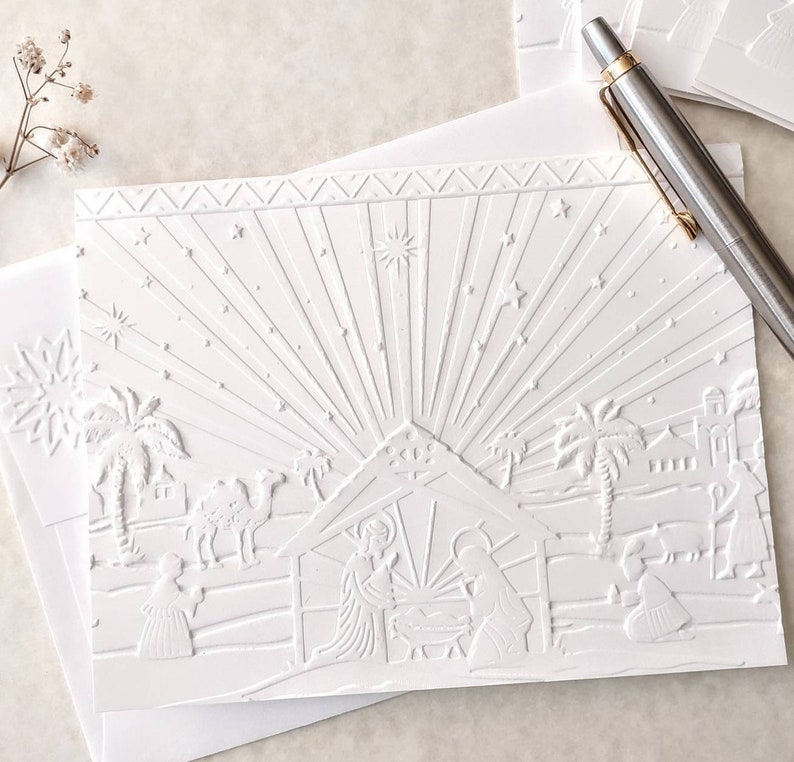 May include: White embossed Christmas card featuring a nativity scene with a starburst design. The card depicts figures, palm trees, camels, and a stable. A silver pen rests on the card, alongside a white envelope and a sprig of white flowers.