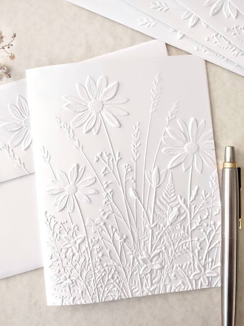 May include: White embossed card with a design of white flowers and foliage. The card is blank and can be used for a variety of occasions.