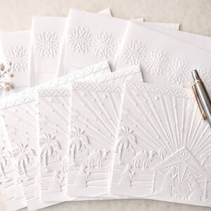 May include: A stack of white embossed Christmas cards. The cards feature a detailed nativity scene with palm trees, camels, and figures. Some cards have a snowflake design. A silver pen rests on the cards.