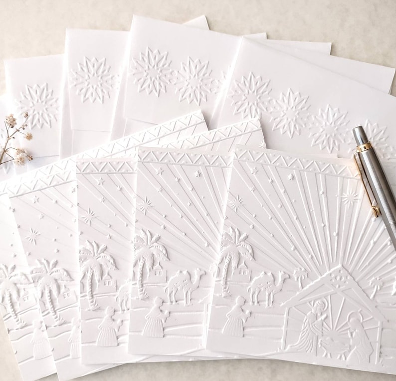May include: A stack of white embossed Christmas cards. The cards feature a detailed nativity scene with palm trees, camels, and figures. Some cards have a snowflake design. A silver pen rests on the cards.