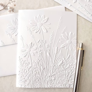 May include: White embossed card with a floral design. The card features a variety of flowers and leaves, including daisies, ferns, and grasses. The design is raised and textured, giving the card a three-dimensional look.