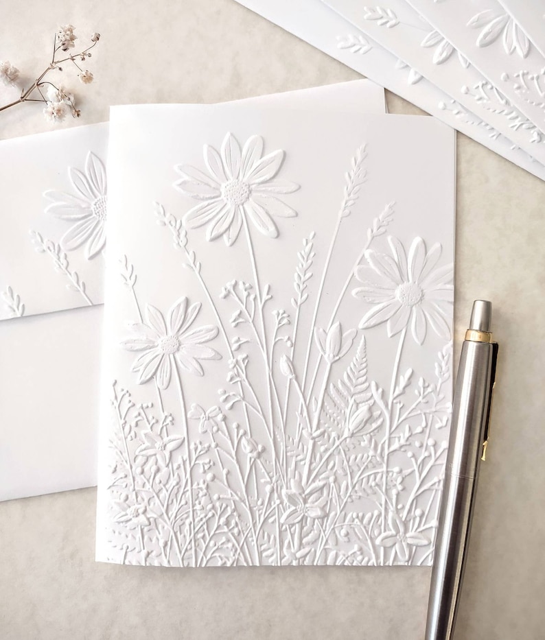 May include: White embossed card with a floral design. The card features a variety of flowers and leaves, including daisies, ferns, and grasses. The design is raised and textured, giving the card a three-dimensional look.