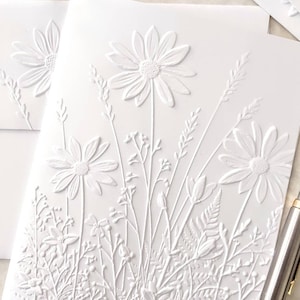 May include: White embossed card with a raised design of white flowers and foliage. The flowers are daisies with a central circle and multiple petals.