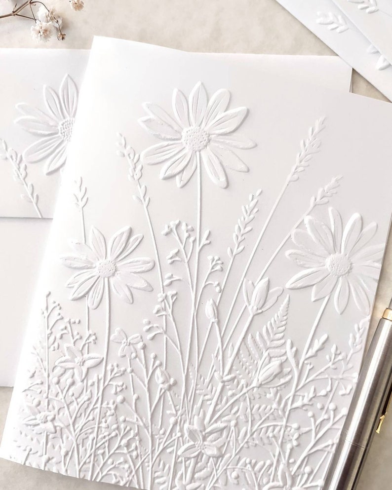 May include: White embossed card with a raised design of white flowers and foliage. The flowers are daisies with a central circle and multiple petals.