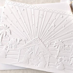 May include: A white embossed Christmas card featuring a nativity scene. The card depicts the holy family, palm trees, camels, and stars. The design includes a radiant burst of light emanating from the stable, with a decorative border at the top.