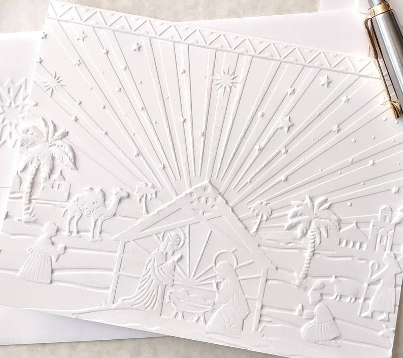May include: A white embossed Christmas card featuring a nativity scene. The card depicts the holy family, palm trees, camels, and stars. The design includes a radiant burst of light emanating from the stable, with a decorative border at the top.