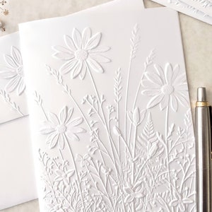 May include: White embossed card with a design of white flowers and foliage. The card is blank and can be used for a variety of occasions.