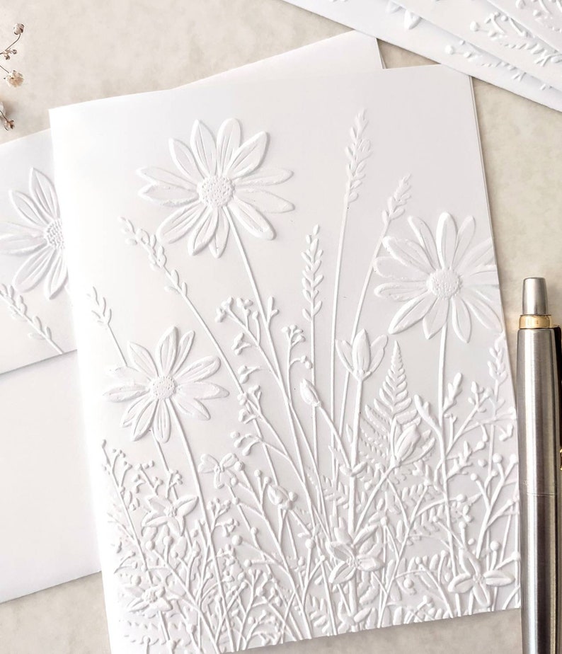 May include: White embossed card with a design of white flowers and foliage. The card is blank and can be used for a variety of occasions.