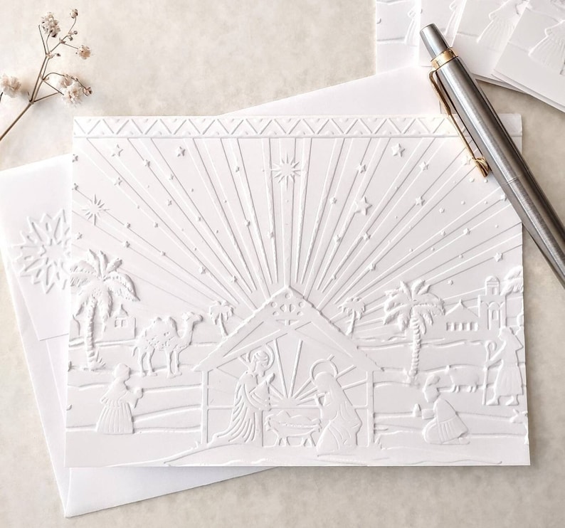 May include: White embossed Christmas cards featuring a nativity scene with a starburst design. The cards depict a stable, figures, and palm trees. Includes envelopes and a pen.
