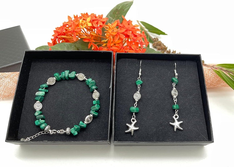 May include: A jewelry set featuring a bracelet and earrings, presented in black boxes. The bracelet and earrings are adorned with green malachite stones and silver-toned spiral and starfish charms. The set is displayed against a backdrop of orange flowers.