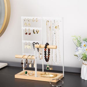 May include: A white metal and wood jewelry organizer displaying various earrings, necklaces, and bracelets. The organizer has multiple tiers for hanging jewelry and a wooden base. The jewelry includes gold, silver, and beaded pieces.