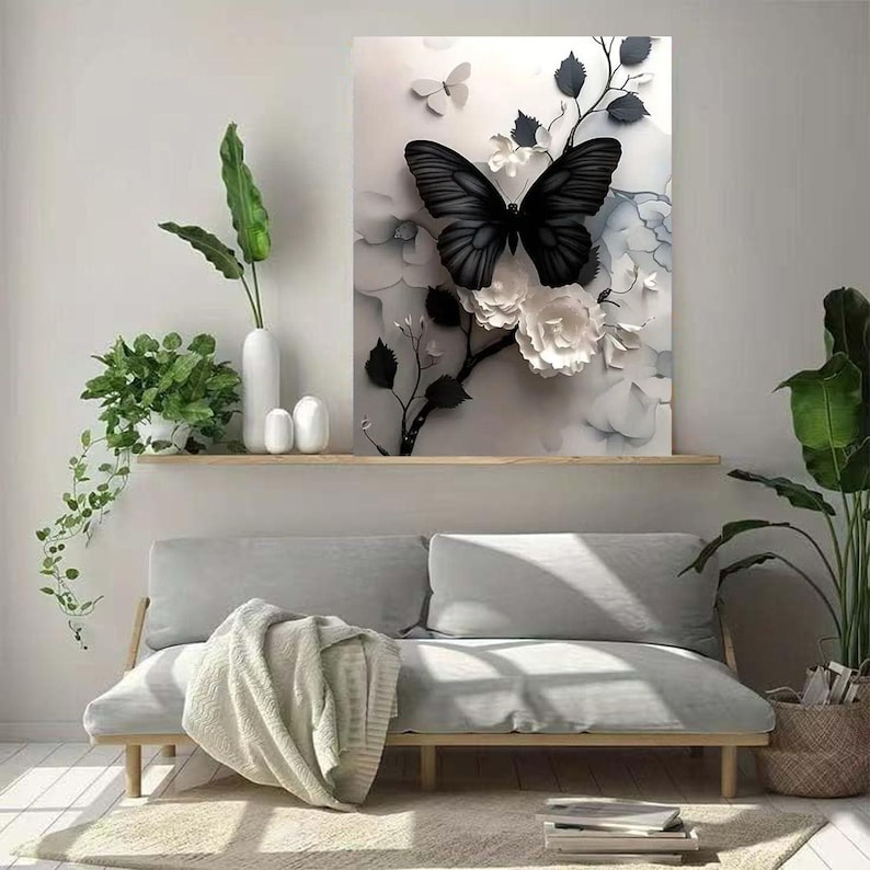 Op de afbeelding: Een ingelijst canvas kunstwerk met een grote zwarte vlinder met gedetailleerde vleugels, tegen een achtergrond van witte bloemen en bladeren. Het kunstwerk wordt boven een lichtgekleurde bank tentoongesteld, wat de inrichting van de kamer verbetert.