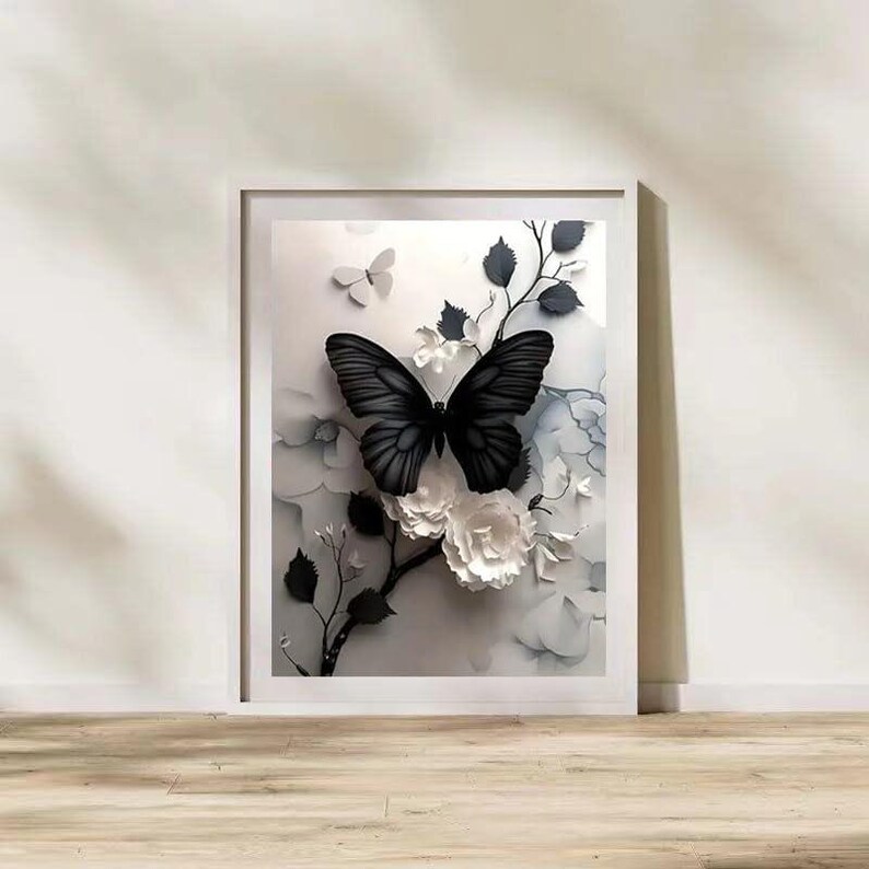 Op de afbeelding: Een ingelijst kunstwerk met een zwarte vlinder met gedetailleerde vleugels, zittend op witte papieren bloemen en takken. Het kunstwerk heeft een 3D-effect, met een zachte, neutrale achtergrond. De lijst is wit en het kunstwerk wordt op een houten oppervlak tentoongesteld.