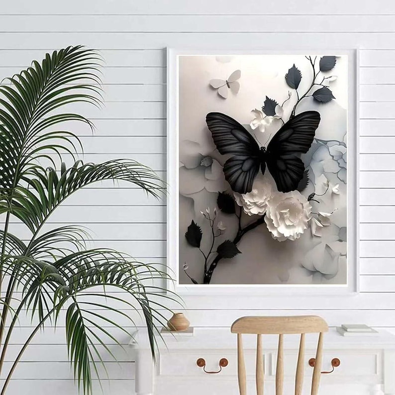 Op de afbeelding: Een ingelijst kunstwerk met een zwarte vlinder met gedetailleerde vleugelpatronen, tegen een achtergrond van witte bloemen en grijs blad. Het kunstwerk zit in een witte lijst, wat een opvallend contrast cre&euml;ert.