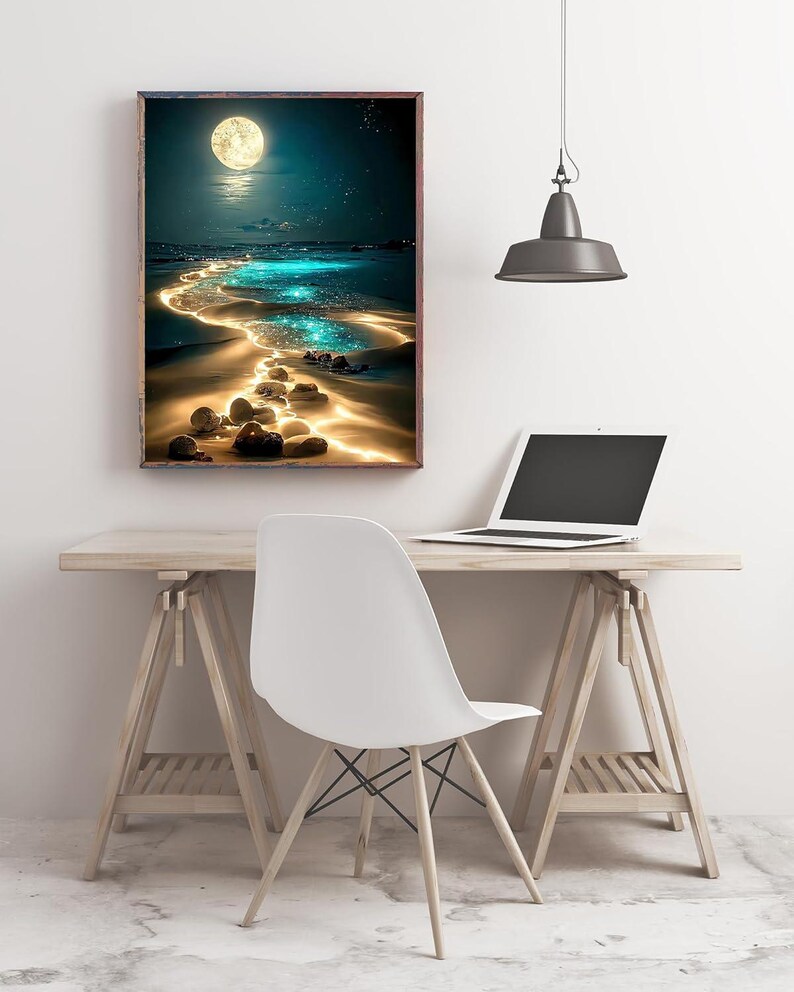 May include: A framed art print depicts a nighttime beach scene with a full moon, glowing water, and a path of illuminated stones. The artwork is displayed on a wooden desk with a white chair and a laptop.