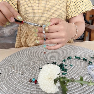 May include: A person is using pliers to assemble a gemstone bracelet. The bracelet features a variety of colorful stones, including turquoise, red, and black. A white flower and other jewelry components are also visible.