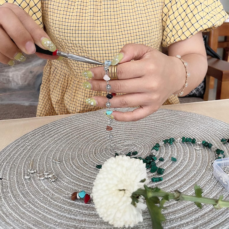 May include: A person is using pliers to assemble a gemstone bracelet. The bracelet features a variety of colorful stones, including turquoise, red, and black. A white flower and other jewelry components are also visible.