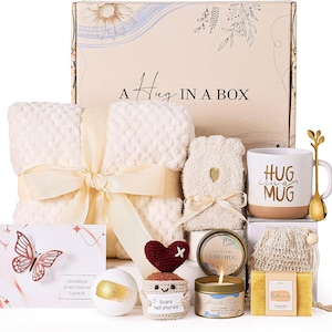 May include: A gift set featuring a cream-colored textured blanket with a ribbon, a mug labeled HUG IN A MUG with a gold spoon, a candle labeled A BIG HUG, a crochet heart figure, a bath bomb, a greeting card with a butterfly, and oatmeal soap.