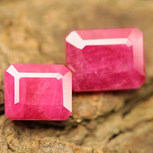 May include: Two rectangular, faceted, pink gemstones with white accents. The gems are displayed on a textured, brown surface, showcasing their vibrant colour and cut. These gems are likely for jewellery making.