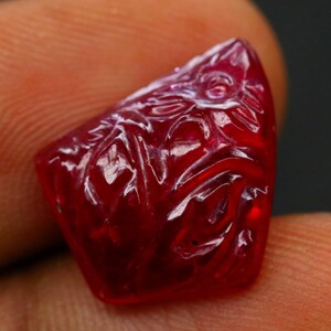 Könnte beinhalten: Ein geschnitzter, tiefroter Edelstein mit aufwendigen floralen oder blattartigen Mustern. Die polierte Oberfläche reflektiert das Licht und hebt die Details der Schnitzerei hervor. Der Edelstein wird zwischen den Fingern gehalten, um seine Größe und Farbe zu zeigen.