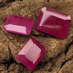 May include: Three faceted, rectangular-cut rubies in varying sizes. The gemstones are a deep, vibrant pinkish-red colour, with a polished, reflective surface. They rest on a textured, brown bark background.