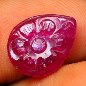 May include: A close-up of a carved, teardrop-shaped ruby gemstone. The gemstone is a deep, vibrant red with intricate floral carvings. The carvings are visible on the surface of the gemstone, adding texture and detail to the design.