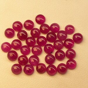 May include: A collection of round, polished ruby gemstones. The rubies are a deep, rich red color and have a smooth, glossy surface. They are scattered across a light beige surface.