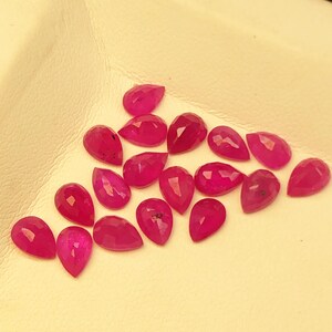 May include: A collection of faceted, teardrop-shaped gemstones in a vibrant magenta hue. The gems are arranged on a light-colored surface, showcasing their polished surfaces and internal clarity. The gemstones are likely for jewelry making.