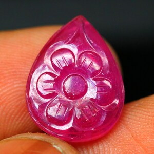 Könnte beinhalten: Ein tropfenförmiger, geschnitzter Rubin-Edelstein. Der Edelstein hat eine leuchtende rosa-rote Farbe und weist ein detailliertes Blumendesign auf, das in seine Oberfläche geschnitzt ist. Die Schnitzerei ist auf dem Stein zentriert.