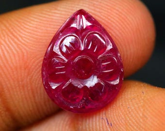May include: A teardrop-shaped, carved ruby gemstone. The deep red stone features a floral design carved into its surface. The gemstone is held between two fingers, showcasing its intricate details and vibrant colour.