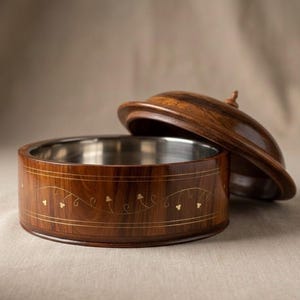 Puede incluir: Un cuenco de madera con tapa, con interior de acero inoxidable. El exterior es de color marrón oscuro con un motivo floral dorado decorativo. La tapa está ligeramente entreabierta, revelando el contenido del cuenco.