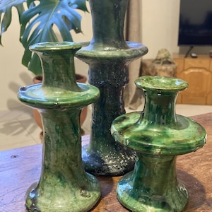 May include: Three handcrafted ceramic candle holders in varying sizes. The holders are glazed in shades of green, with a textured, organic appearance. The tallest holder is approximately 10 inches tall, while the smallest is about 5 inches.