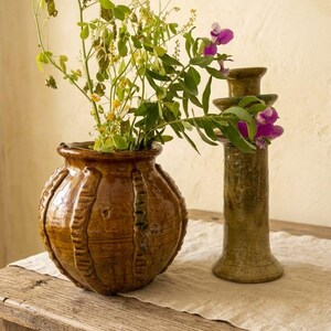 May include: A round, brown glazed ceramic pot with vertical ribbed detailing and a matching tall, slender candlestick holder. The pot contains a bouquet of green stems with small yellow flowers and purple blossoms extending outward.