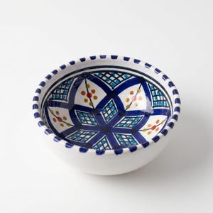 May include: A round ceramic bowl with a white base and a hand-painted interior pattern featuring dark blue geometric lines, a grid motif, and small orange and yellow floral accents. The rim is decorated with a repeating dark blue striped pattern.
