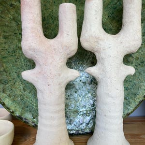 May include: Two off-white ceramic candle holders with a textured surface. Each holder has a U-shaped top and a flared base. The background features a green ceramic plate and a wooden surface.