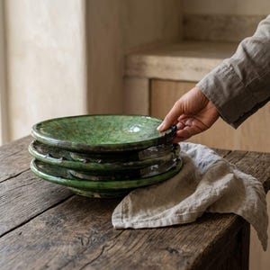 Puede incluir: Una pila de tres platos redondos de cerámica con un esmalte verde brillante y moteado. Los platos presentan bordes irregulares formados a mano y un centro profundo en forma de cuenco. La superficie del esmalte muestra variaciones de color.