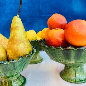 Puede incluir: Tres cuencos de cerámica verde llenos de fruta fresca. Peras, limones y naranjas se exhiben en los cuencos. Los cuencos tienen un borde festoneado y se asientan sobre una base de pedestal. El fondo es azul liso.